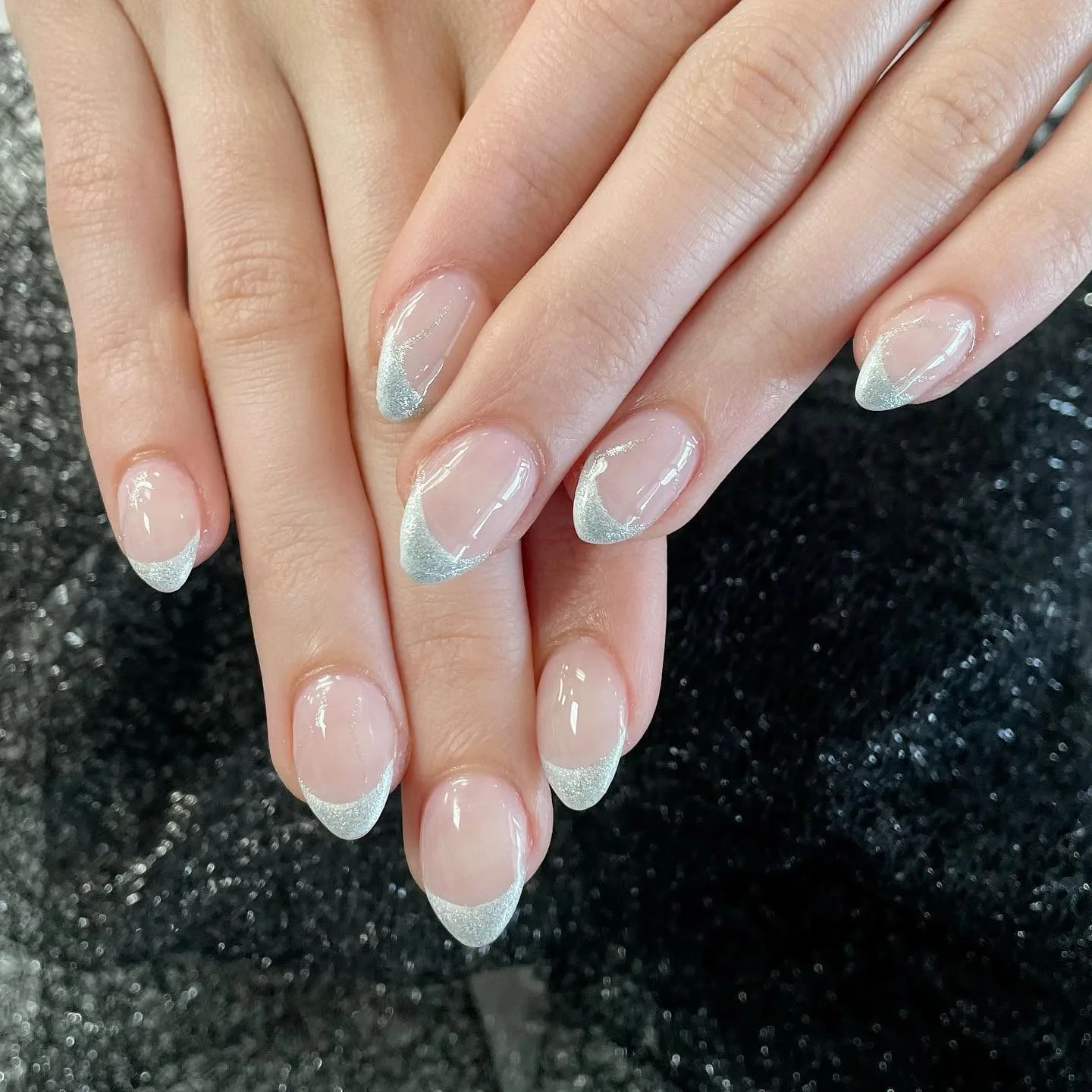 Almond nails with a sheer pink base and silver glitter French tips, against black sparkly fabric