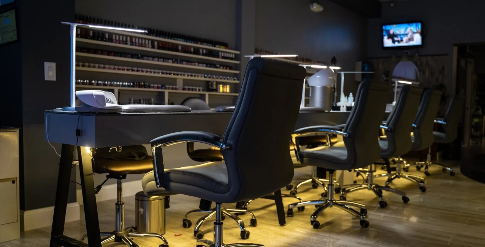A row of manicure tables with desk lamps and black chairs, in front of shelves of nail polish